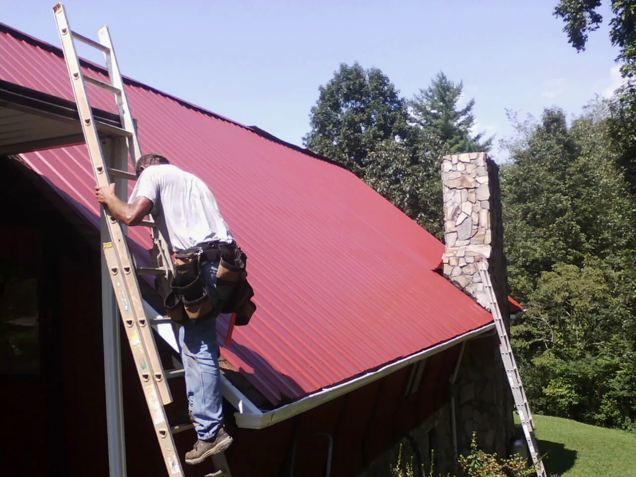 Roofer on a ladder beside a red metal roof and stone chimney — Soffit & Fascia Repair by Roofing Brothers in Tillman